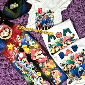 May include: A collection of Super Mario-themed apparel, including a bucket hat, overalls, and several white t-shirts. The overalls and shirts feature colorful graphics of Mario, Luigi, and other characters. The text "MOM" and "DAD" are on two shirts.