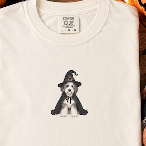 Könnte beinhalten: Ein cremefarbenes T-Shirt mit einer zentralen Grafik eines flauschigen Hundes, der einen schwarzen Hexenhut und einen passenden schwarzen Umhang trägt. Das T-Shirt hat ein Comfort Colors Markenetikett am Innenkragen mit der Größenangabe L G G.