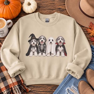 May include: A beige crewneck sweatshirt featuring a graphic print of four fluffy dogs in Halloween costumes. The dogs are dressed as a witch, a skeleton, a ghost, and a vampire. The sweatshirt has long sleeves, a ribbed collar, and ribbed cuffs.