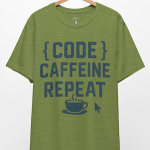 May include: Green t-shirt with the text "{CODE} CAFFEINE REPEAT" in blue. The design includes a coffee cup and a computer mouse cursor. The shirt is hanging on a wooden hanger.