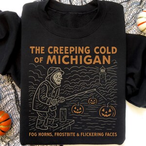 May include: Black sweatshirt with a graphic design featuring a fisherman, lighthouse, and jack-o'-lanterns. The text reads "The Creeping Cold of Michigan" and "Fog Horns, Frostbite & Flickering Faces". Decorative pumpkins are in the background.