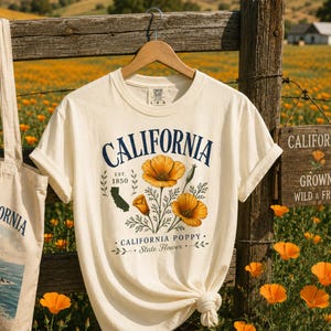 Puede incluir: Camiseta color crema con "CALIFORNIA" en azul marino, con un dise&ntilde;o floral de amapolas de California naranjas. Tambi&eacute;n se ven una bolsa de mano a juego y un letrero de madera con el mismo tema. La camiseta est&aacute; colgada en una valla de madera en un campo de flores naranjas.