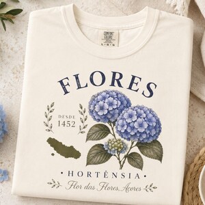 May include: A cream-colored t-shirt featuring a graphic print of blue hydrangea flowers, green leaves, and a map silhouette. The text reads FLORES, DESDE 1452, HORTÊNSIA, and Flor das Flores, Acores. The shirt has a crew neckline and a Comfort Colors label.