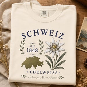 May include: A cream-colored t-shirt featuring a graphic with the text SCHWEIZ, SEIT 1848, EDELWEISS, and Schweizer Nationalblume. The design includes a green map of Switzerland, a detailed illustration of an edelweiss flower, and leaf accents.