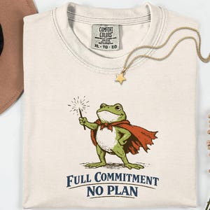 May include: Cream-colored t-shirt featuring a cartoon frog in a red cape holding a sparkler. The text "FULL COMMITMENT NO PLAN" is printed below the frog. A gold necklace with a star pendant and a Comfort Colors tag are also visible.