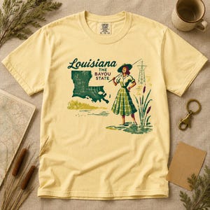 May include: A light yellow t-shirt featuring a graphic of Louisiana with the text "Louisiana The Bayou State". The graphic includes a woman fishing, a map of Louisiana, and a power line tower. The shirt is a Comfort Colors XL.