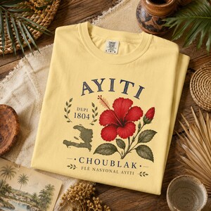 May include: A pale yellow crew neck t-shirt featuring a graphic print with the text AYITI, DEPI 1804, and CHOUBLAK FL&Egrave; NASYONAL AYITI. The design includes a red hibiscus flower, a green map of Haiti, and decorative leaf sprigs in a vintage style.