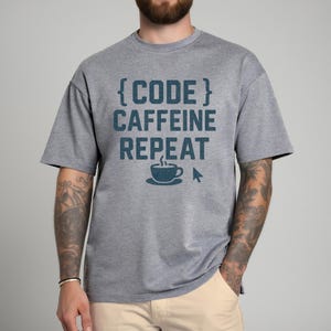 Pode incluir: Camiseta cinza com o texto "{CODE} CAFFEINE REPEAT" em verde-azulado. O design inclui uma x&iacute;cara de caf&eacute; e um cursor de computador. A camiseta &eacute; de estilo casual, adequada para o uso di&aacute;rio.