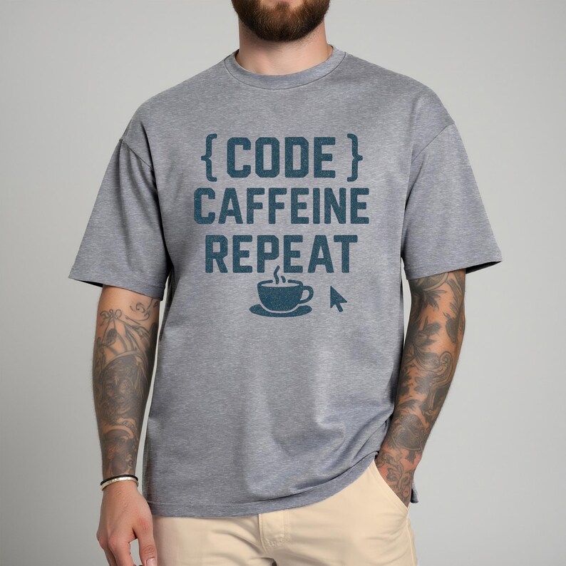 Pode incluir: Camiseta cinza com o texto "{CODE} CAFFEINE REPEAT" em verde-azulado. O design inclui uma x&iacute;cara de caf&eacute; e um cursor de computador. A camiseta &eacute; de estilo casual, adequada para o uso di&aacute;rio.