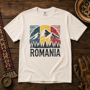 May include: An off-white short-sleeve t-shirt featuring a distressed graphic of the Romanian flag colors in a circular design. The print includes mountain peaks, a forest of pine trees, and the word ROMANIA in bold, dark blue capital letters.