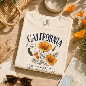Puede incluir: Camiseta color crema con la palabra "CALIFORNIA" en azul marino, con un gr&aacute;fico de amapolas de California y el texto "CALIFORNIA POPPY - State Flower". La camiseta incluye el texto "EST. 1850" y una silueta de California.