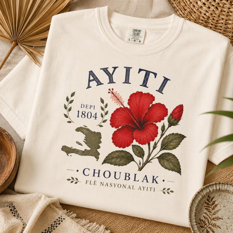 May include: An off-white t-shirt featuring a graphic print with the text AYITI, DEPI 1804, CHOUBLAK, and FL&Egrave; NASYONAL AYITI. The design includes a red hibiscus flower with green leaves, a bud, and a dark green silhouette map of the country of Haiti.