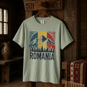 May include: A light sage green short-sleeve t-shirt featuring a distressed graphic print of the Romanian flag colors in a circular design. The graphic includes a mountain silhouette, a forest of pine trees, and the word ROMANIA in bold text.