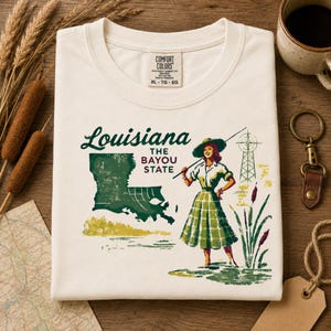 May include: Cream-colored t-shirt with a graphic of Louisiana, the Bayou State. The design features a woman in a green plaid skirt, a map of Louisiana, and the text "Louisiana The Bayou State".