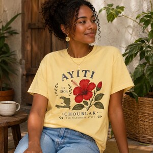 May include: A pale yellow short-sleeve t-shirt featuring a graphic print of a red hibiscus flower, a map of Haiti, and the text AYITI, DEPI 1804, CHOUBLAK, and FLE NASYONAL AYITI. The shirt is paired with blue denim jeans.