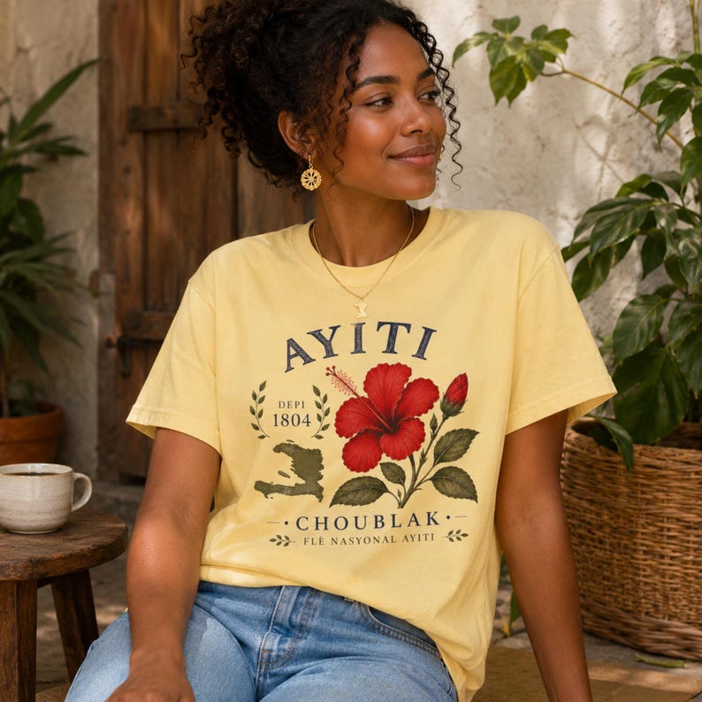 May include: A pale yellow short-sleeve t-shirt featuring a graphic print of a red hibiscus flower, a map of Haiti, and the text AYITI, DEPI 1804, CHOUBLAK, and FLE NASYONAL AYITI. The shirt is paired with blue denim jeans.