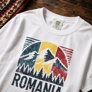 May include: A white short-sleeve t-shirt featuring a distressed graphic of the Romanian flag colors in a circular design. The print includes silhouettes of mountains and pine trees with the word ROMANIA in bold dark blue capital letters.