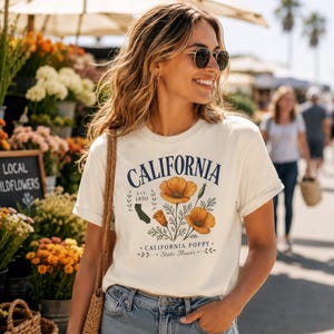 Puede incluir: Camiseta color crema con la palabra "CALIFORNIA" en texto arqueado azul marino, y una ilustraci&oacute;n de amapolas de California. La camiseta tambi&eacute;n incluye el texto "EST. 1850" y "CALIFORNIA POPPY. State Flower."