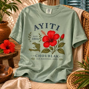 May include: A sage green short-sleeve t-shirt featuring a graphic print with the text AYITI, DEPI 1804, CHOUBLAK, and FL&Egrave; NASYONAL AYITI. The design includes a large red hibiscus flower, green leaves, and a map outline of Haiti in olive green.