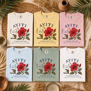 May include: Six short-sleeved t-shirts in cream, yellow, pink, light blue, sage green, and white. Each shirt features a graphic of a red hibiscus flower, the text AYITI, DEPI 1804, CHOUBLAK, FLE NASYONAL AYITI, and a map outline of Haiti.