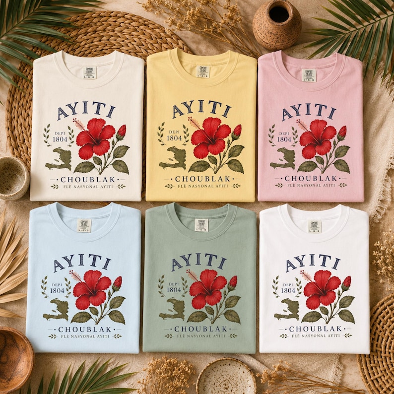 May include: Six short-sleeved t-shirts in cream, yellow, pink, light blue, sage green, and white. Each shirt features a graphic of a red hibiscus flower, the text AYITI, DEPI 1804, CHOUBLAK, FLE NASYONAL AYITI, and a map outline of Haiti.