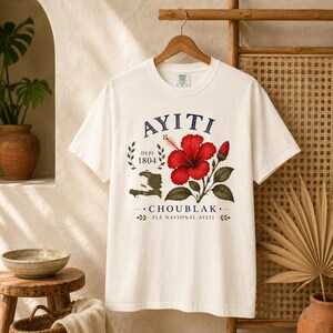 May include: An off-white short-sleeve t-shirt featuring a graphic print with the text AYITI, DEPI 1804, and CHOUBLAK FLE NASYONAL AYITI. The design includes a red hibiscus flower, a green map of Haiti, and decorative leaf accents.