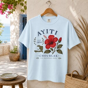 May include: A light blue short-sleeve t-shirt featuring a graphic print with the text AYITI, DEPI 1804, and CHOUBLAK FLE NASYONAL AYITI. The design includes a red hibiscus flower, a green leaf branch, and a map silhouette of Haiti in dark green.