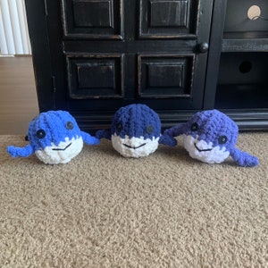 May include: Three handmade crochet whale plush toys in shades of blue and white. Each whale has button eyes and a smiling face. The toys are arranged on a carpeted surface.