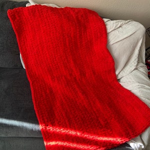 May include: A vibrant red, hand-knitted blanket draped over a gray sofa. The blanket's texture is visible, with thick, plush yarn creating a cozy appearance. The blanket is folded, and the sunlight casts shadows across the surface.