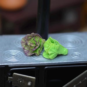 Pode incluir: Fogão preto em miniatura com tampo prateado, com dois pequenos alimentos esculpidos: uma flor roxa e verde e uma alface verde brilhante. O fogão tem uma porta aberta e uma chaminé preta.