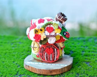 May include: A crocheted mushroom house with a red door, hearts, and other colorful details. The house is sitting on a wooden base.