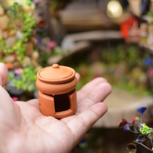 Puede incluir: Un pequeño tiesto de arcilla de color terracota con tapa, diseñado para parecer una estufa o horno en miniatura. El tiesto tiene una pequeña abertura en la parte delantera y la tapa es ligeramente abovedada. El tiesto está siendo sostenido en la mano de una persona.