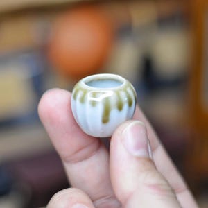 May include: A small, round, white ceramic pot with a light blue interior and a decorative olive green glaze dripping down the sides. The pot is held in a person's hand.