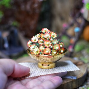以下が含まれることがあります： 小さな白い花とオレンジの花でいっぱいのミニチュアの金のボウル。ボウルは小さな織られたマットの上に置かれています。