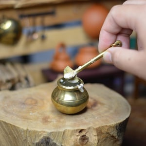 Puede incluir: Una pequeña olla de latón y una cuchara, llenas de una sustancia blanca, están sobre una superficie de madera. Una mano sostiene la cuchara, sacando el contenido. La escena sugiere una cocina en miniatura o un tema relacionado con la comida.