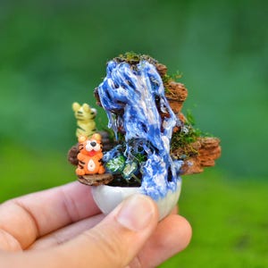 May include: Miniature waterfall scene featuring a blue waterfall, mossy rocks, and two small tiger figurines. The scene is set in a small white ceramic pot, creating a whimsical miniature landscape.
