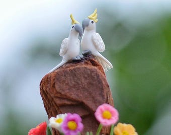 May include: Two white cockatoo figurines with yellow crests and gray beaks perched on a brown rock. The base features colorful flowers in pink, yellow, and red. The background is blurred.