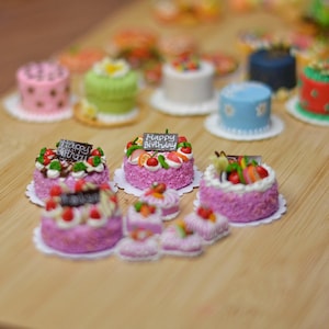 May include: A collection of miniature cakes in various colors, including pink, green, blue, and red, with white frosting and decorative toppings. Some cakes have "Happy Birthday" signs. The cakes are arranged on a light brown wooden surface.
