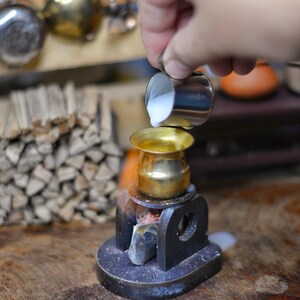 Puede incluir: Una escena de cocina en miniatura con una pequeña olla de latón sobre una estufa negra. Una mano vierte líquido de una pequeña jarra de metal en la olla. Una pila de leña y otras herramientas de cocina en miniatura están en el fondo.