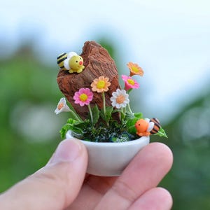 Puede incluir: Una escena de jardín en miniatura en una maceta de cerámica blanca, con flores coloridas, follaje verde y pequeñas figuras de una abeja y un pez payaso. El arreglo incluye un trozo de madera.