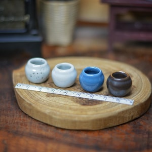 Puede incluir: Cuatro pequeñas macetas de cerámica en varios colores: gris claro moteado, blanco, azul y marrón. Están dispuestas sobre un disco de madera con una cinta métrica. Las macetas tienen aproximadamente 2,5 cm de diámetro, una colección de cerámica en miniatura.