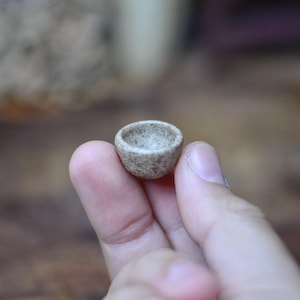 Könnte beinhalten: Eine kleine, handgefertigte Keramikschale mit einer gesprenkelten, hellbraunen Oberfläche. Die Miniatur-Schale wird in der Hand einer Person gehalten, was ihre zarte Größe und rustikale Textur hervorhebt. Das Innere der Schale ist heller.