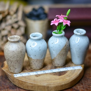 May include: Four small ceramic vases in varying shades of beige and blue-grey sit on a wooden base. One vase holds pink flowers. A ruler is placed in front of the vases. The vases have a speckled design.
