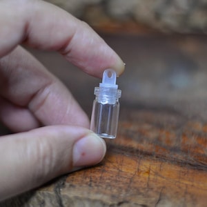 Puede incluir: Un pequeño vial de vidrio transparente con un tapón de plástico blanco. El vial se sostiene entre los dedos contra un fondo de madera borroso. El tapón tiene un pequeño lazo en la parte superior.
