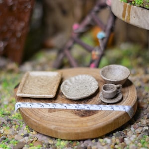 Puede incluir: Juego de té de cerámica en miniatura con plato cuadrado, plato redondo, cuenco, taza y platillo. El juego es de color marrón claro y se encuentra sobre una superficie de madera. Una cinta métrica se coloca junto al juego.