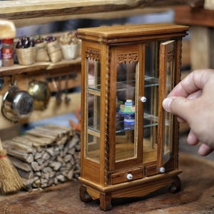 Może przedstawiać: Miniaturowa drewniana szafka ze szklanymi drzwiami i półkami, prezentująca małe przedmioty w środku. Szafka jest brązowa z białymi gałkami i jest częścią miniaturowej sceny kuchennej. Ręka otwiera drzwi szafki.