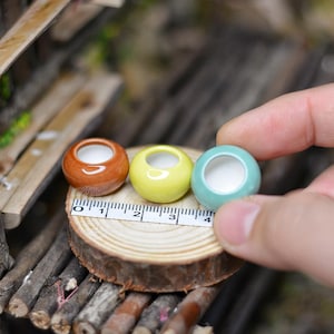 May include: Three miniature ceramic pots in brown, yellow, and teal, each with a white interior. The pots are arranged on a small wooden slice with a ruler, suggesting a craft or miniature project. The pots are approximately 2.5 cm in diameter.
