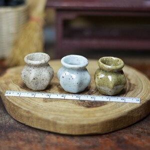 May include: Three small ceramic vases in speckled beige, light blue, and olive green sit on a wooden slice. The vases have a rounded shape with a narrow opening. A ruler is placed in front of the vases, showing measurements in centimetres.