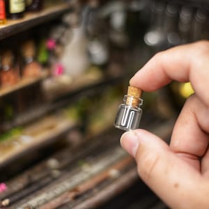 May include: A small, clear glass bottle with a cork stopper. The bottle is being held by a hand.