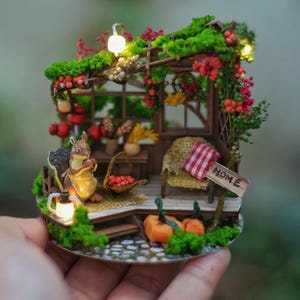 May include: Miniature diorama of a cozy garden scene with a squirrel, pumpkins, and a sign that says "HOME". The scene includes a small chair with a red and white checkered pillow, a basket of red apples, and decorative flowers.